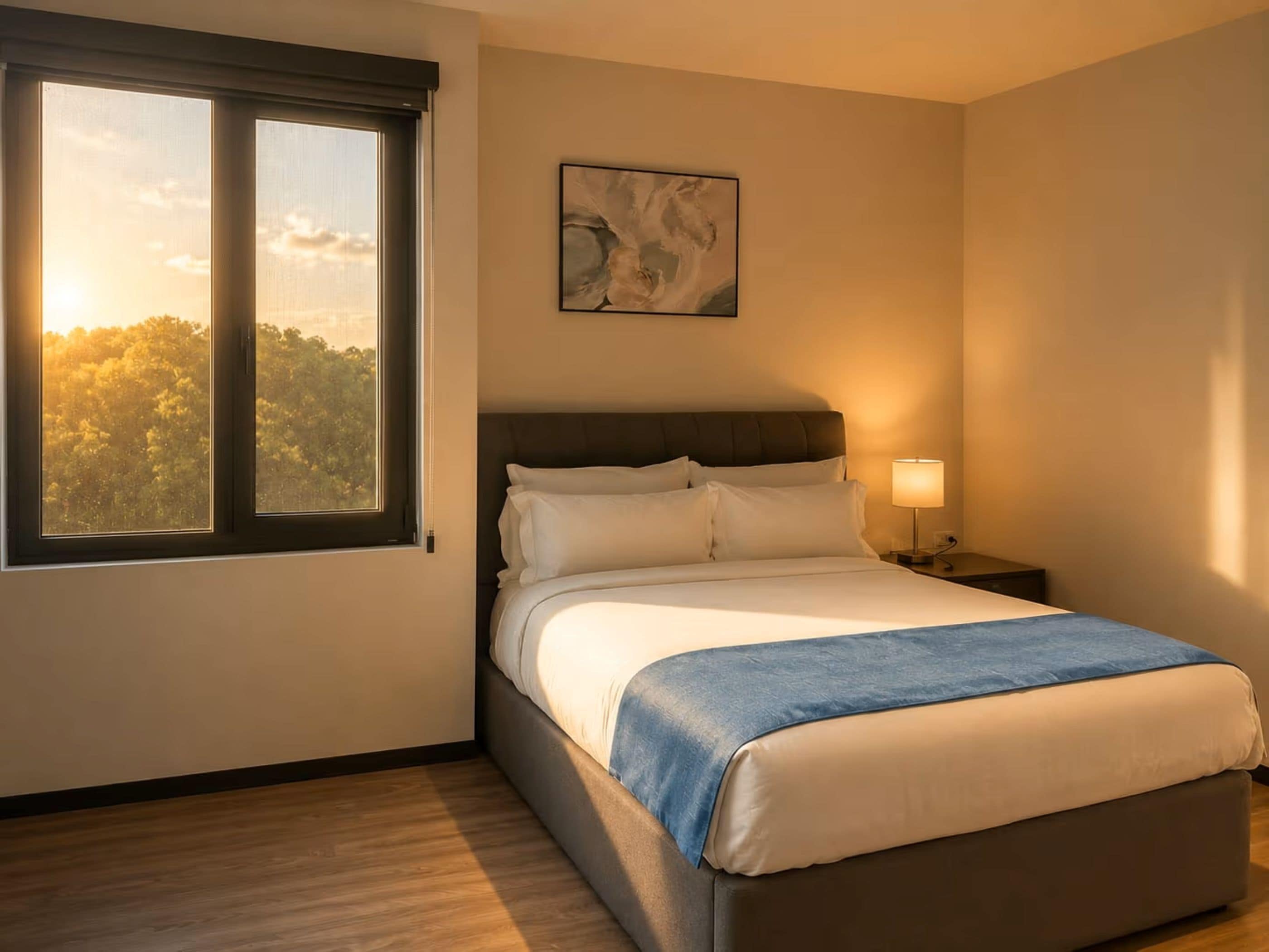 Ein Schlafzimmer mit Bett, Nachttisch, Lampe, Fenster mit Blick auf Bäume und einem Bild an der Wand.