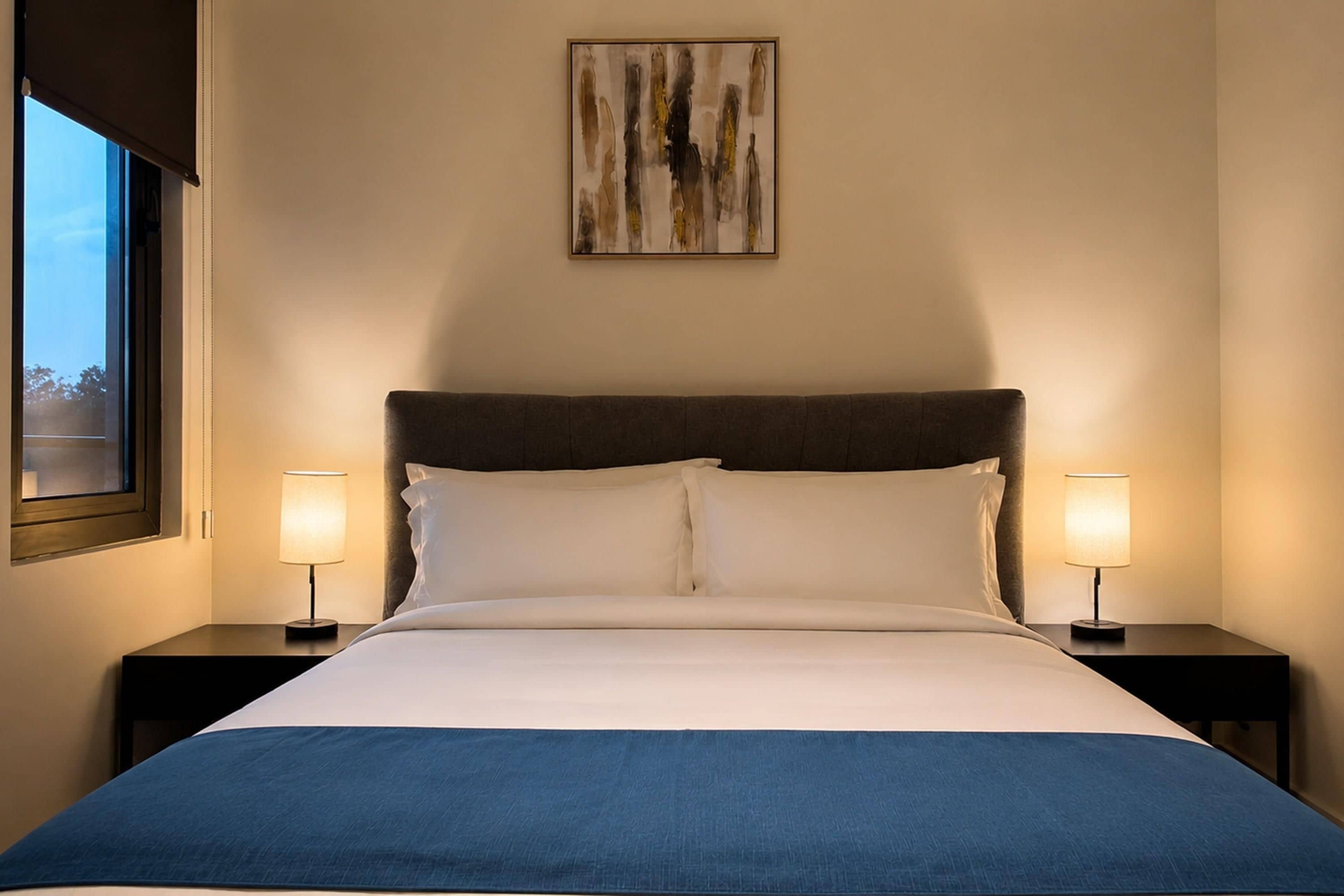 Una habitación de hotel con una cama, dos lámparas de noche, un ventanal y un cuadro en la pared.