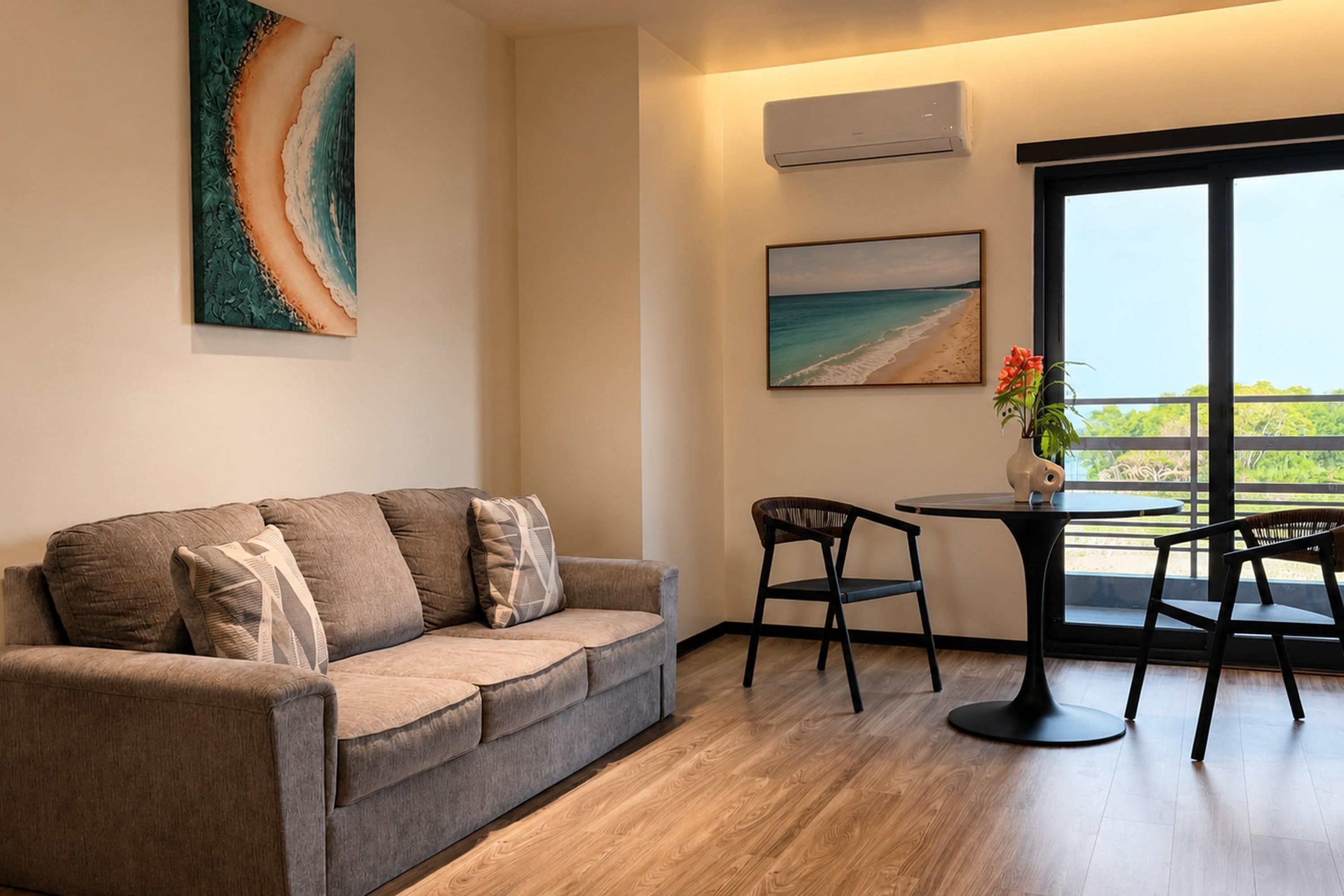 A modern living room with a grey sofa, a small round table, and two chairs. There are two wall-mounted air conditioners and two framed pictures.