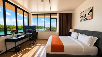 A hotel room with a large window, a bed, a desk with a computer, and a painting on the wall.