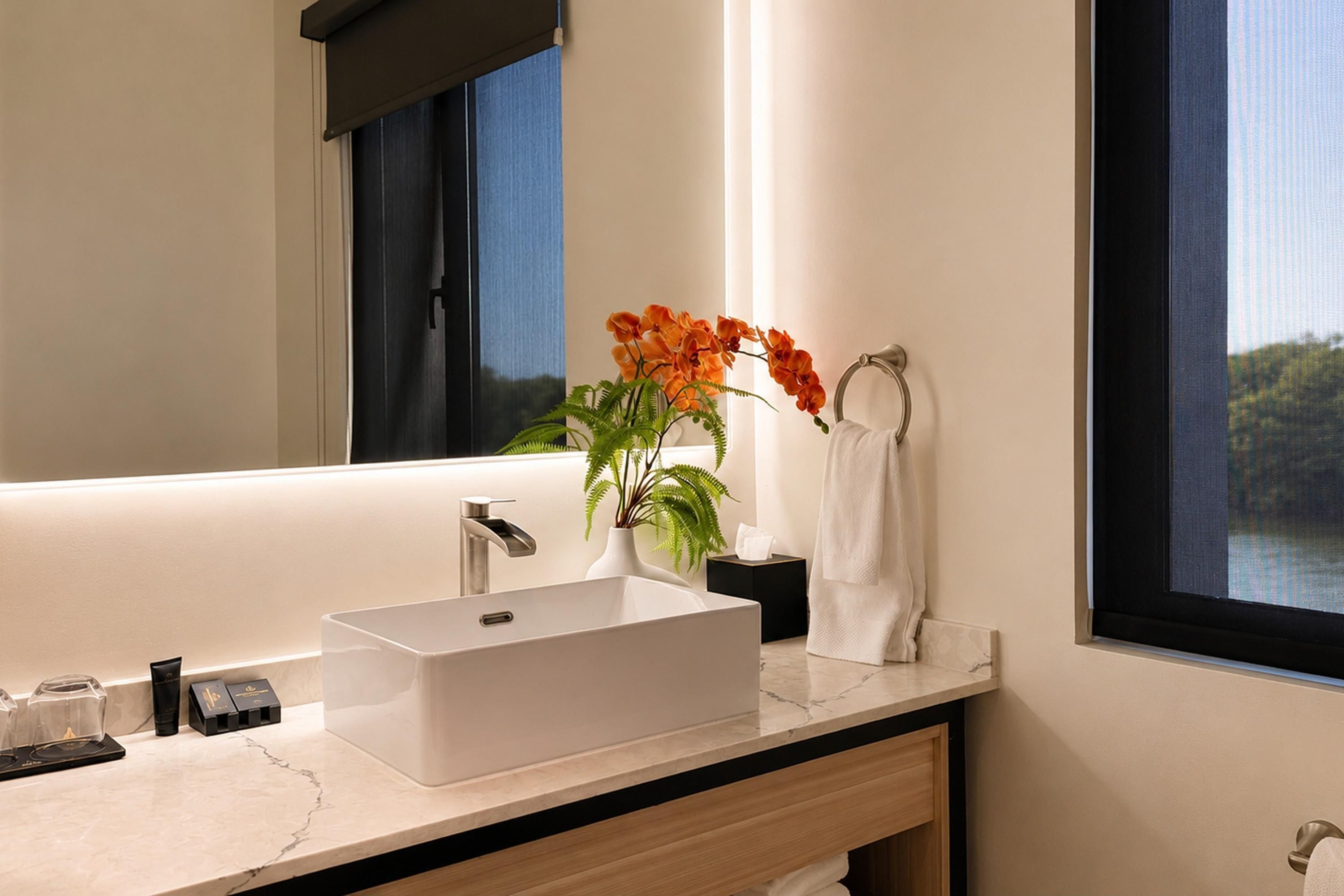 A modern bathroom with a large mirror, a rectangular sink, and a window with a view of greenery.