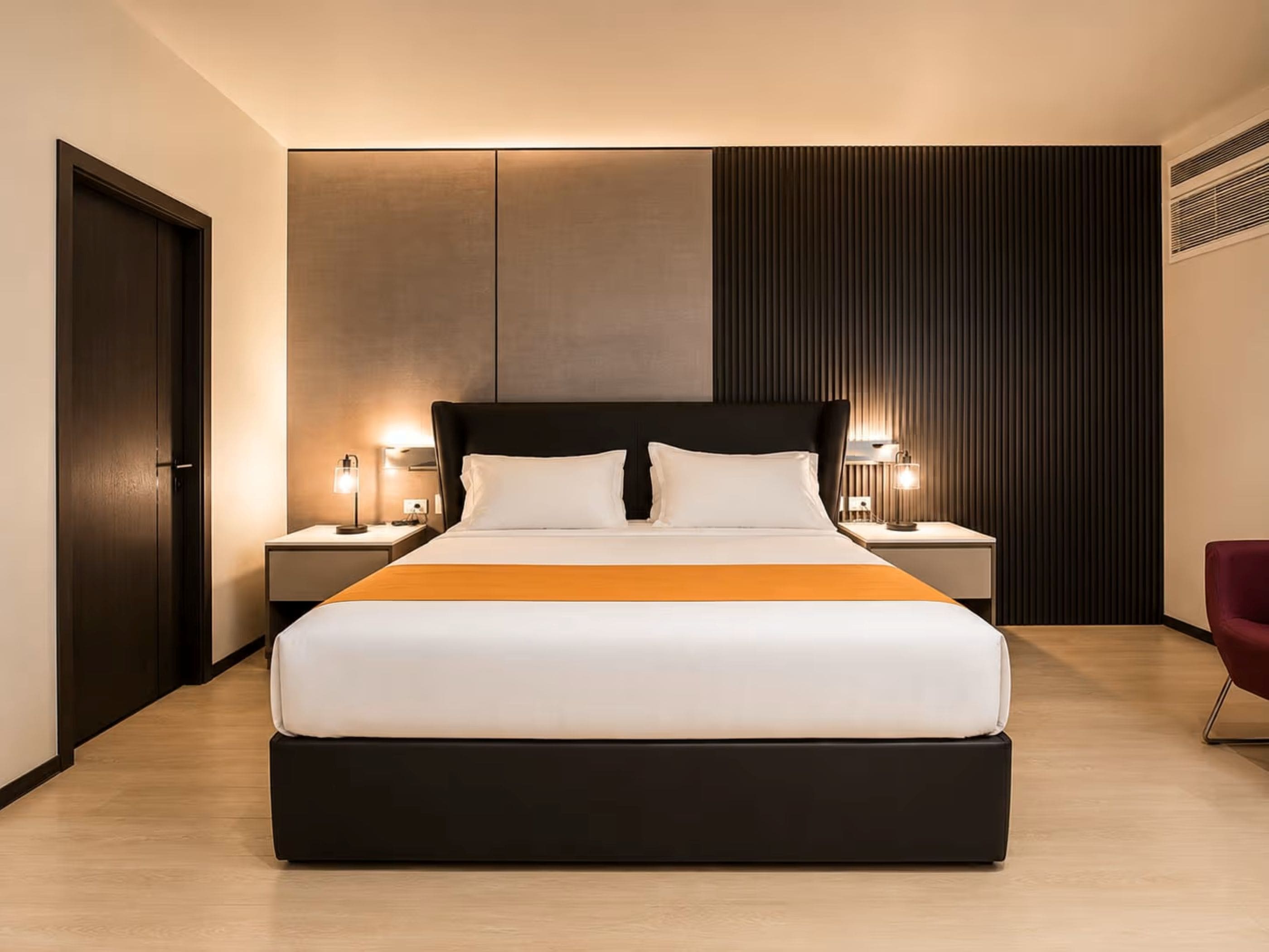 A modern hotel room with a large bed, two bedside tables with lamps, a dark headboard, and a wooden floor.