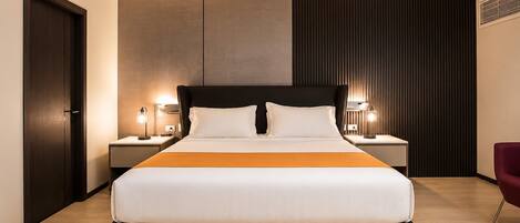 Una habitación de hotel moderna con una cama grande, dos mesitas de noche con lámparas, un cabecero oscuro y suelo de madera.