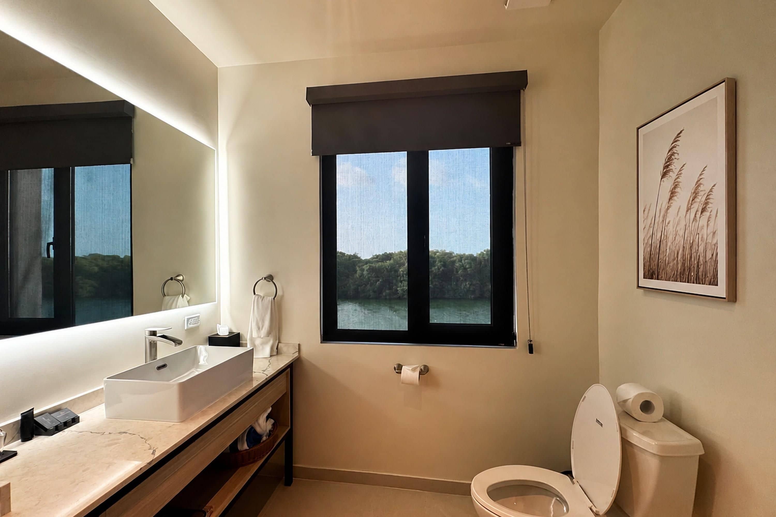 A modern bathroom with a large mirror, a sink, a toilet, and a window with a view of greenery.
