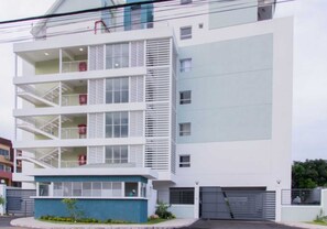 Edifício residencial de vários andares com varandas, garagem e pequena área de entrada com plantas.