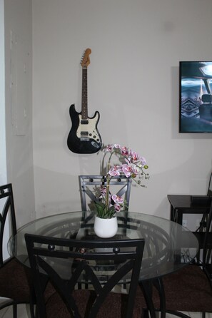 A dining area with a glass table surrounded by chairs, a potted orchid, and a guitar on the wall.