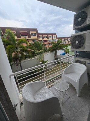 A balcony with two white wicker chairs and a small round table, overlooking a residential area with buildings and palm trees.