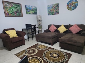 Una sala de estar con un sofá y sillón marrones, un escritorio pequeño con una computadora portátil y un tapete decorativo.