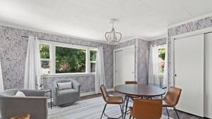 Un coin repas avec une table ronde, quatre chaises et un lustre. Il y a deux fenêtres avec des rideaux, une porte et un mur orné d’un motif.