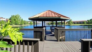 En lusthus med en träveranda som leder ner till en sjö, omgivet av grönska och bostadshus.