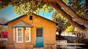 A small, single-story building with a blue door and windows, a brick pathway, and a picnic table under a tree.