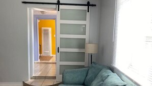 A living room with a green sofa, a sliding glass door, a lamp, and a view of a hallway with a yellow door.