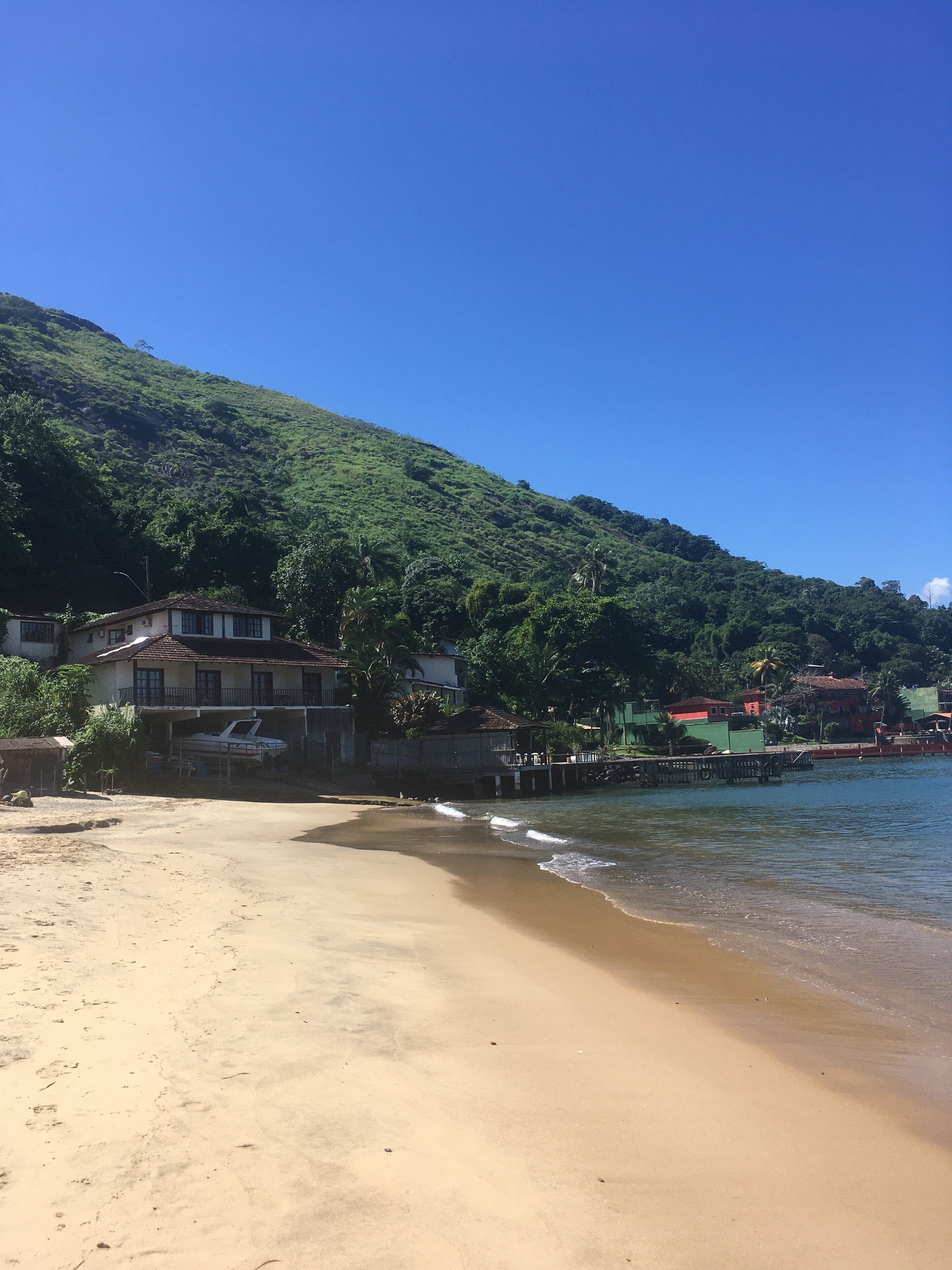 Uma praia com areal, um barco pequeno e casas com uma colina ao fundo.