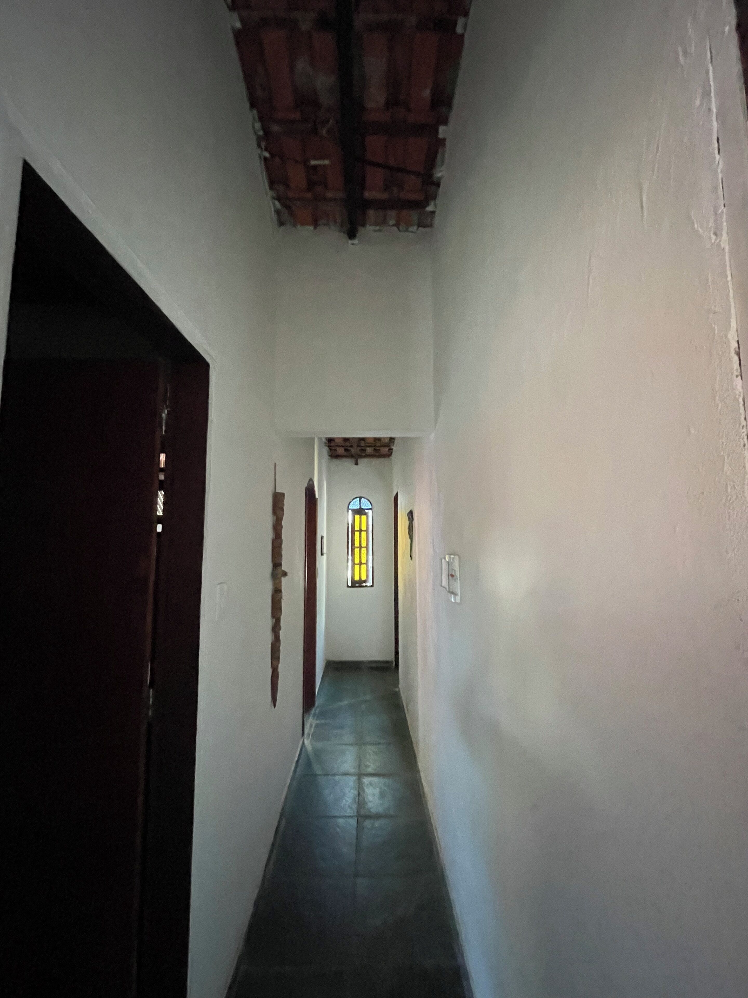 A narrow hallway with a tiled floor, white walls, and a wooden ceiling. There is a door on the left and a window with yellow-tinted glass at the end of the hallway.