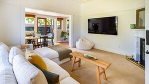A living room with a sofa, a coffee table, and a fireplace. There is a television mounted on the wall and a dining area visible through an open door. - Exclusive 2 Bed Standalone Villa in St Albans (Christchurch)
