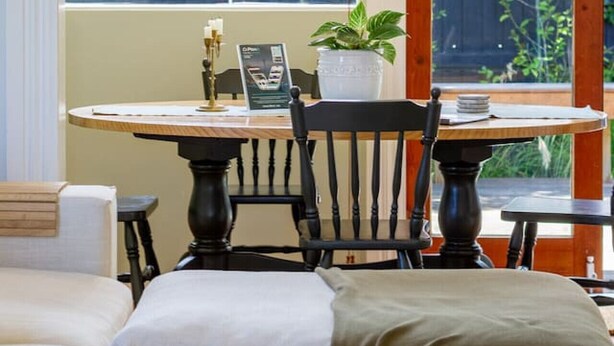 A cozy room with a bed, a dining table with chairs, a small table with a plant, and a window with a view of a fence and trees. - Exclusive 2 Bed Standalone Villa in St Albans (Christchurch)