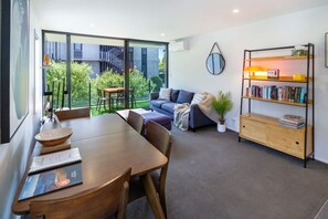 Una sala de estar moderna con comedor, un sofá, una estantería y vistas a un edificio y zonas verdes al exterior.