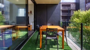 Et moderne udendørs spiseområde med et træbord og stole, omgivet af glasvægge og udsigt til en bygning med balkon.