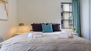 En pænt redt seng med to håndklæder, et natbord med en lampe, og et vindue med udsigt til en bygning.