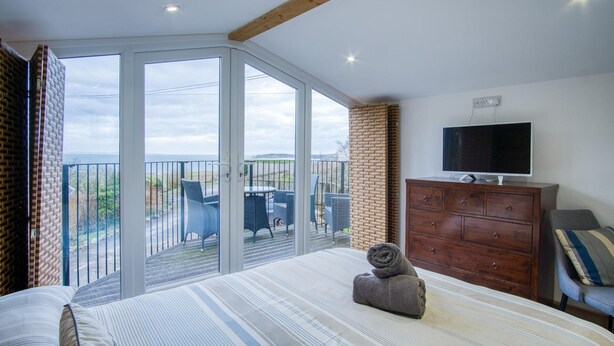 Een slaapkamer met een grote schuifpui naar een balkon, een houten dressoir, een televisie en een bed met gestreepte lakens.