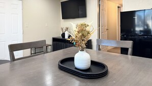 Un coin repas comprenant une table, des chaises, un vase avec des fleurs séchées, un plateau noir, un téléviseur à écran plat et un réfrigérateur.