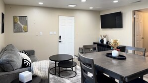 Un salon moderne avec un canapé gris, une table basse ronde, une table à manger avec des chaises, un téléviseur à écran plat fixé au mur et des spots encastrés au plafond.