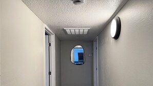 Un couloir avec une bouche d’aération au plafond, un petit miroir rond accroché au mur et une porte au fond.