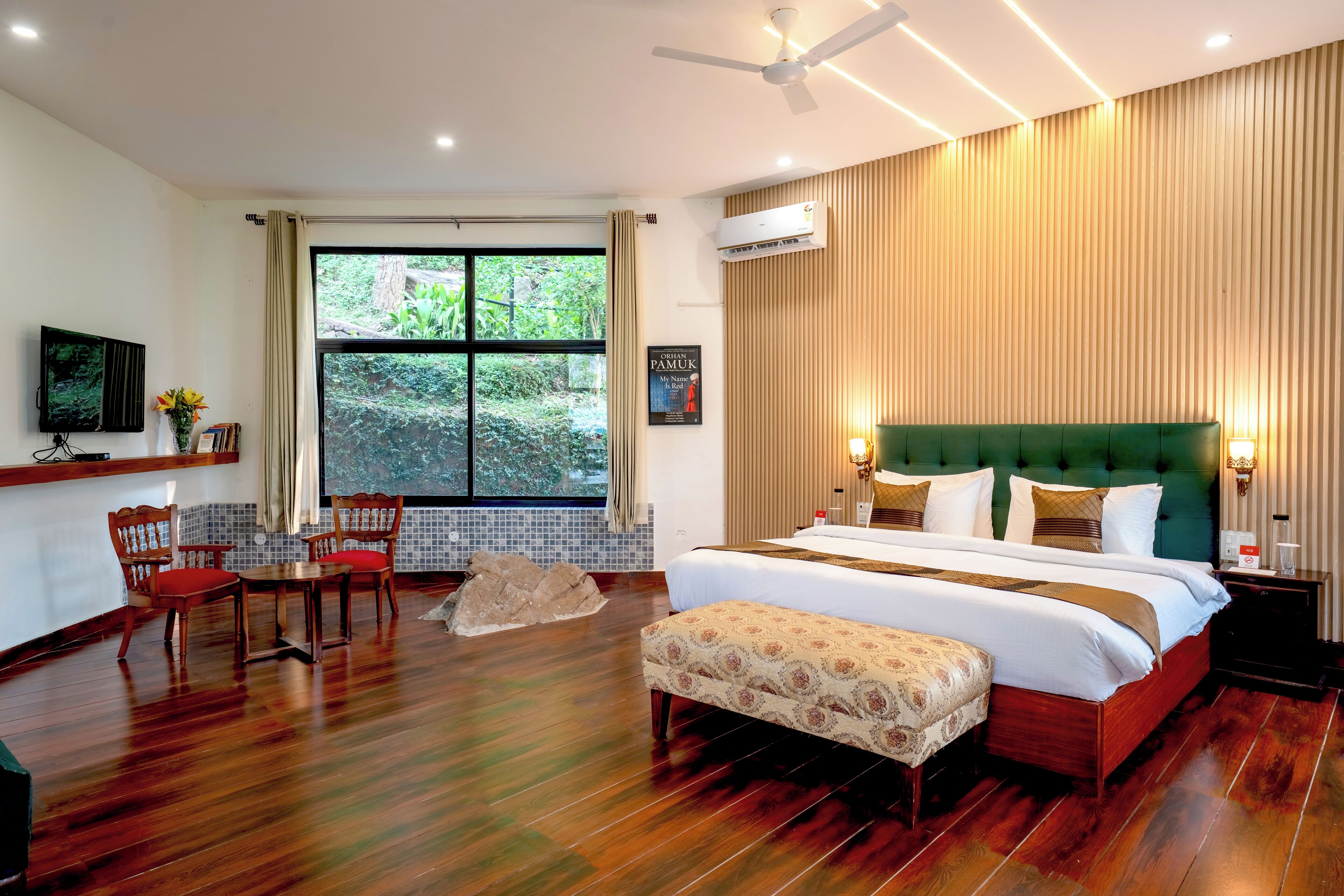 A hotel room with a large bed, a green upholstered headboard, a wooden floor, a dining area with chairs, a small table, and a large window with a view of greenery.