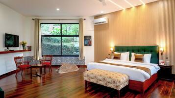 A hotel room with a large bed, a green upholstered headboard, a wooden floor, a dining area with chairs, a small table, and a large window with a view of greenery.