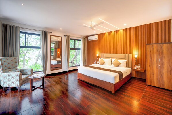A modern hotel room with a large bed, a sitting area with a chair and table, and large windows offering a view of trees.