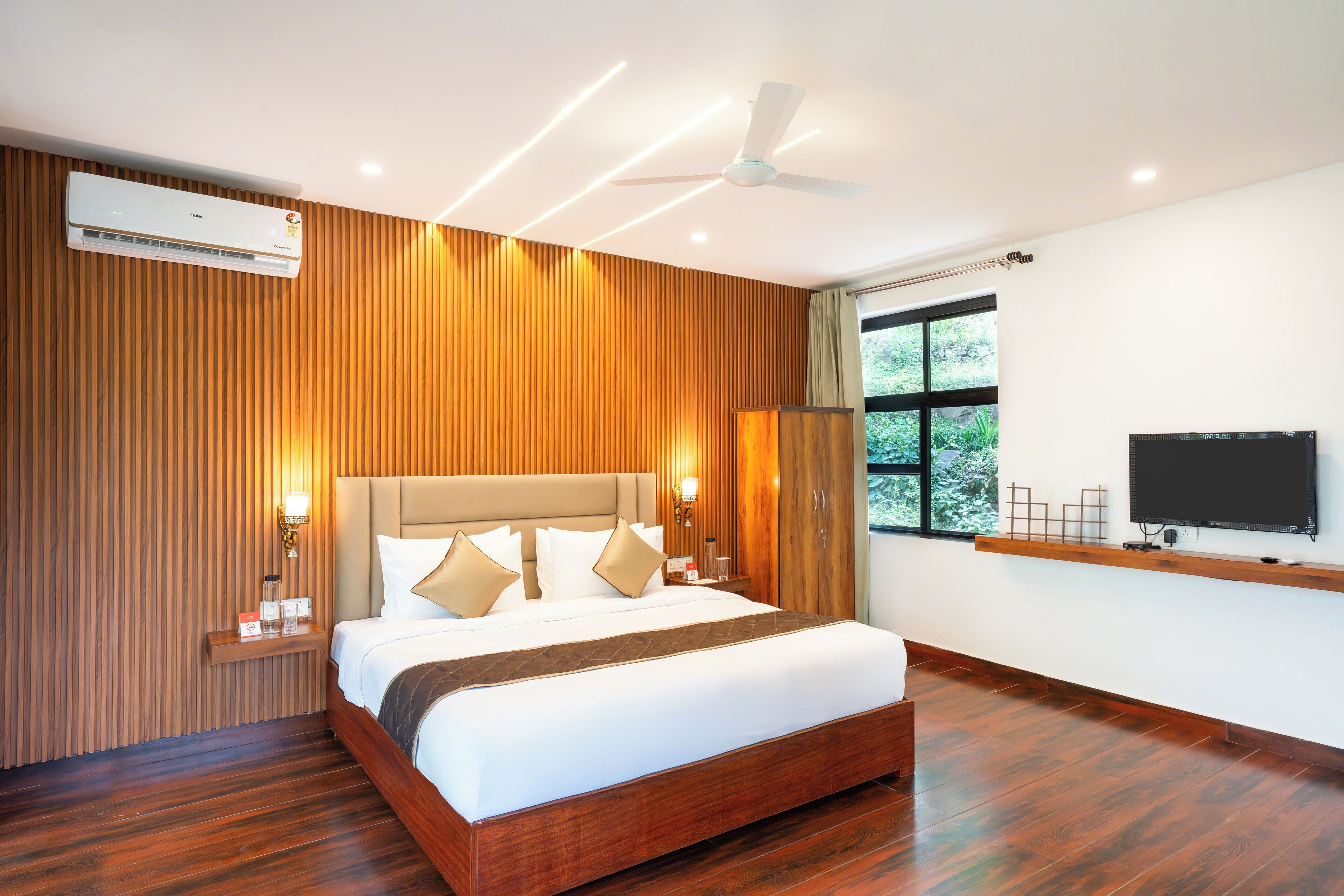 Una habitación de hotel moderna con pared de paneles de madera, una cama grande con ropa de cama blanca, un ventilador de techo, un televisor montado en la pared y una ventana con vistas a la vegetación.