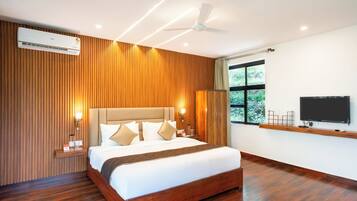 Una habitación de hotel moderna con pared de paneles de madera, una cama grande con ropa de cama blanca, un ventilador de techo, un televisor montado en la pared y una ventana con vistas a la vegetación.