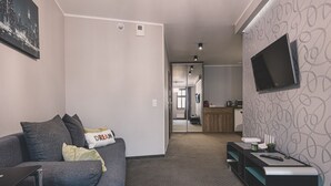 Un salon moderne avec un canapé gris, un téléviseur à écran plat fixé au mur et un coin cuisine à l’arrière-plan.