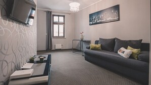 Un salon moderne avec un canapé gris, un téléviseur à écran plat fixé au mur, un tableau représentant un paysage urbain et un lustre.