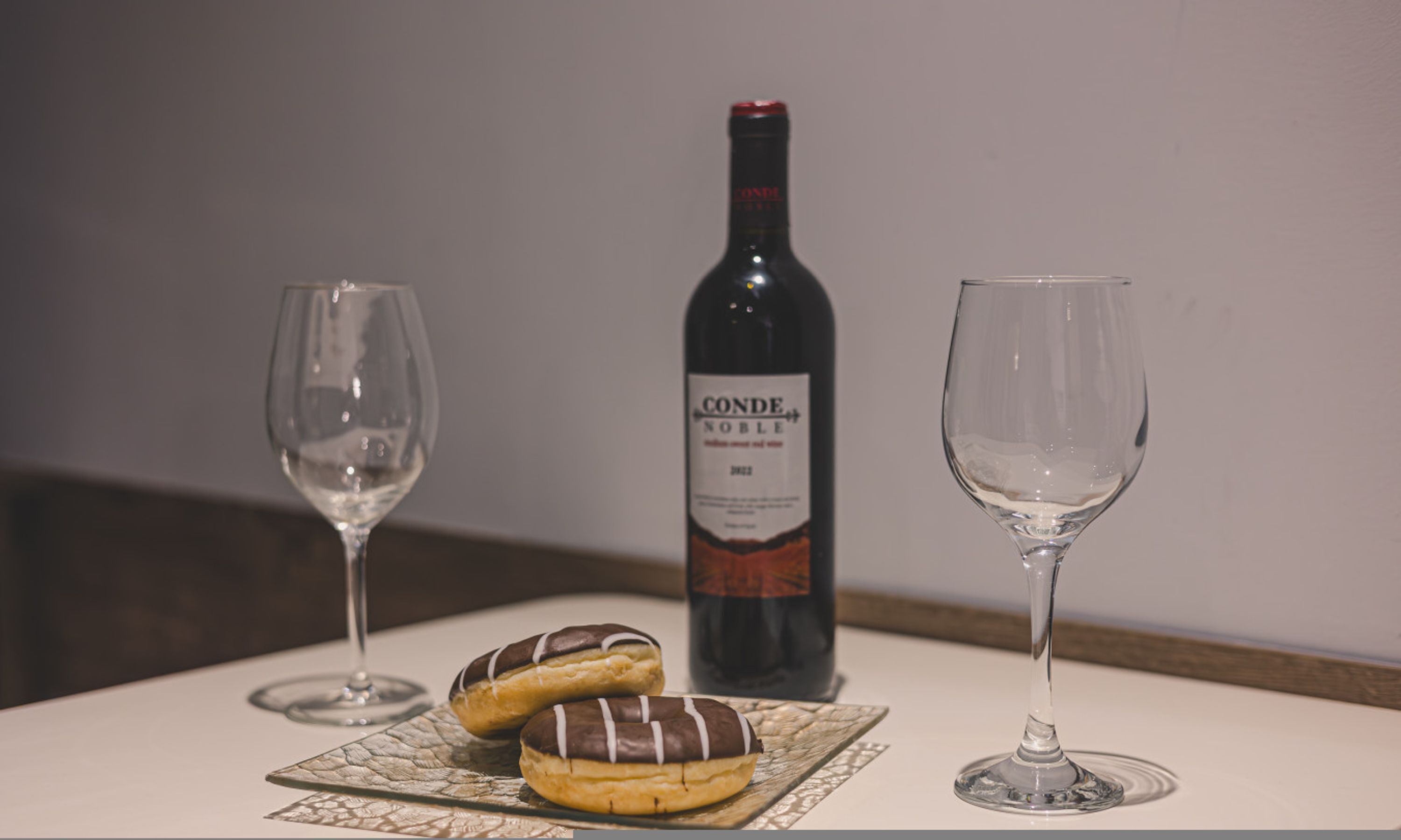 Two wine glasses, a bottle of Conde Noble wine, and two chocolate-covered donuts on a table.