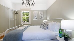 A bedroom with a bed, a window with a view of trees, a chandelier, and a bedside table with a bottle and a plant. - Secret Garden - historic charm meets Bowral vibe (Bowral)