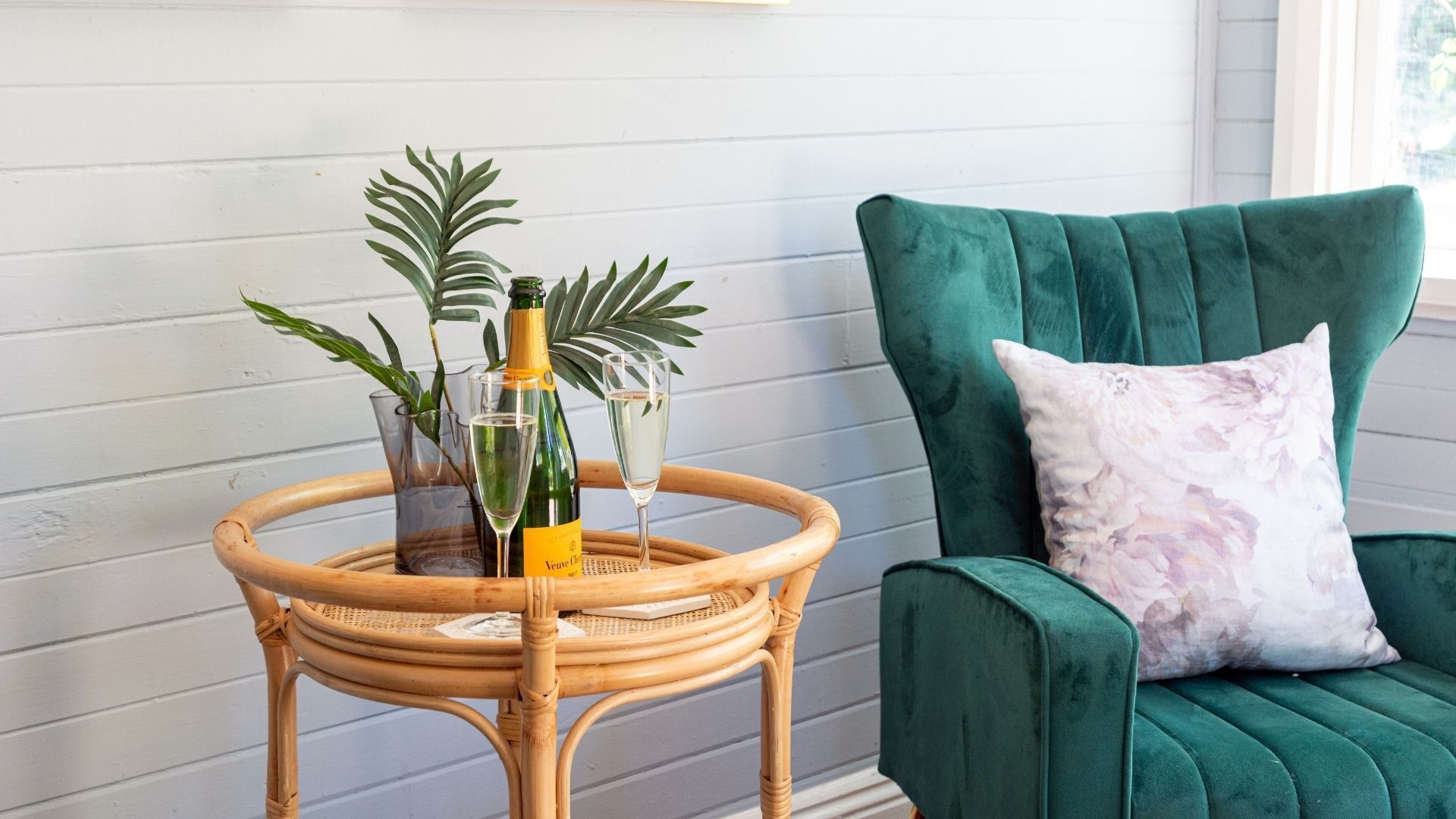 Une petite table d’appoint en osier, sur laquelle se trouvent une bouteille de champagne, des verres et des plantes. Une chaise à l’assise verte, ornée d’un coussin à motifs floraux, est placée à côté.
