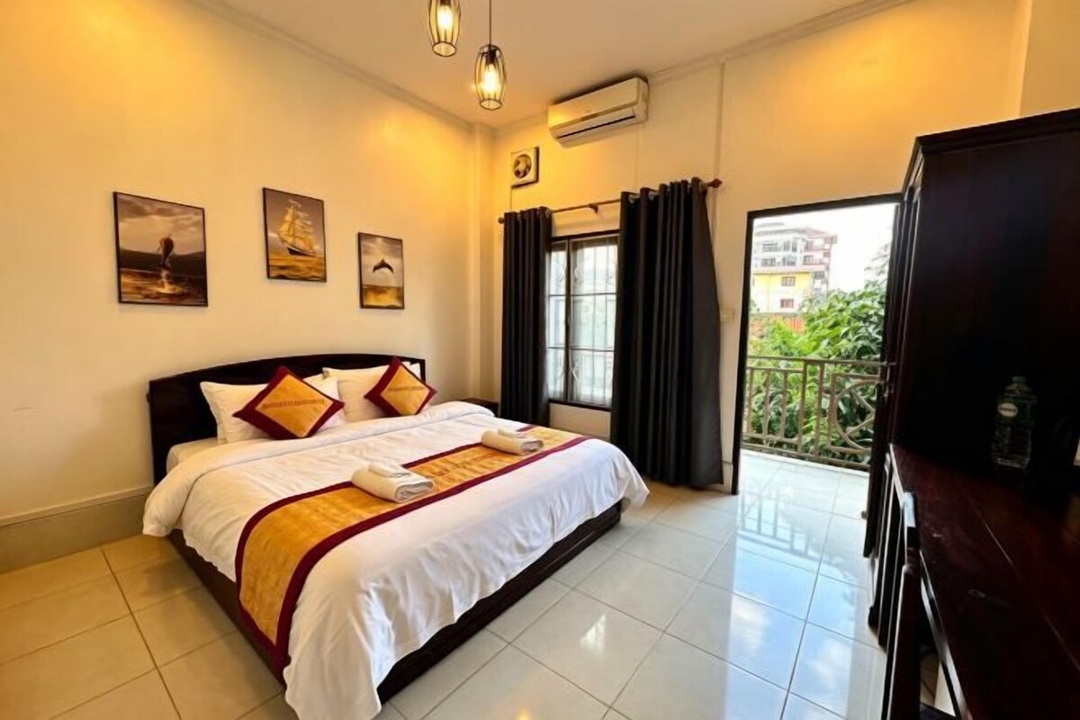 A hotel room with a bed, a flat-screen TV, a balcony with curtains, and artwork on the wall.