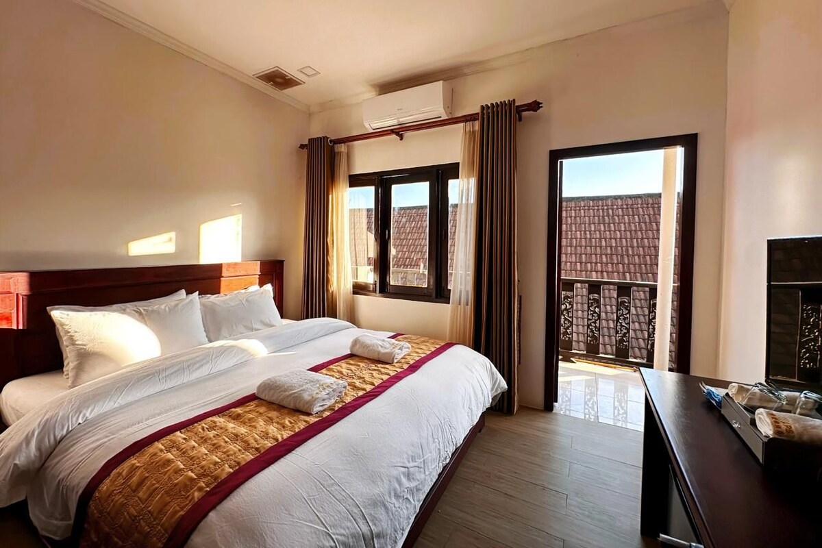 A hotel room with a large bed, a wooden headboard, a bedside table with a mirror, and a view of a building through the window.