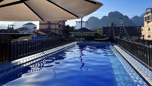 A swimming pool with a tiled edge, a large umbrella, and a view of buildings and mountains in the background. - Vang Vieng Family Boutique Hotel (Vang Vieng)