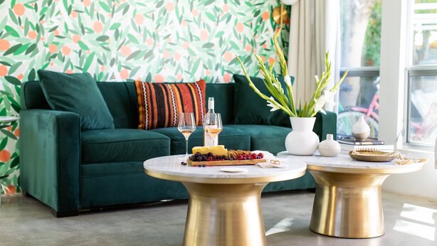 Ein Wohnzimmer mit einem grünen Sofa, zwei goldenen Tischen mit Speisen und Getränken sowie einem gemusterten Wandpapier.