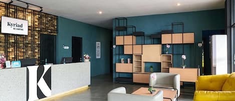 Un área de recepción moderna con un destacado letrero de 'Kyriad', una mesa de recepción, espacios de asiento y una pared decorativa con estantes.