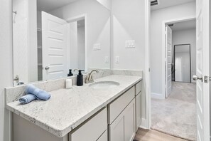 Une salle de bain avec un comptoir blanc, un lavabo et un miroir. On y trouve des serviettes sur le comptoir, ainsi qu’une porte qui donne sur une autre pièce.