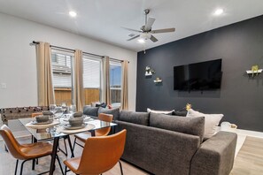 Un salon moderne comprenant un coin repas, un canapé gris, un téléviseur à écran plat fixé au mur et un ventilateur de plafond.