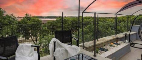 An outdoor terrace with a table and chairs, a view of greenery and water, and a sunset sky.