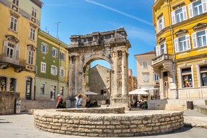 Ein historischer Steinbogen in einem öffentlichen Platz, umgeben von Gebäuden und Außenbereich mit Sitzgelegenheiten.