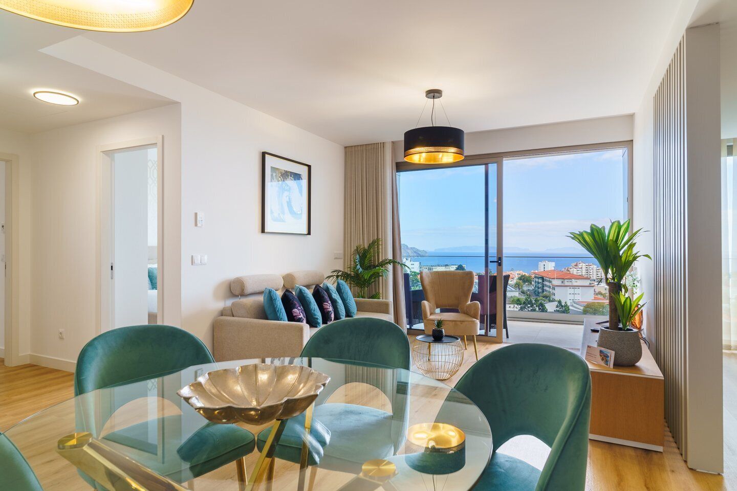 A modern living room with a glass dining table, green chairs, a beige sofa with blue cushions, a large window with a city view, and a hanging light fixture.