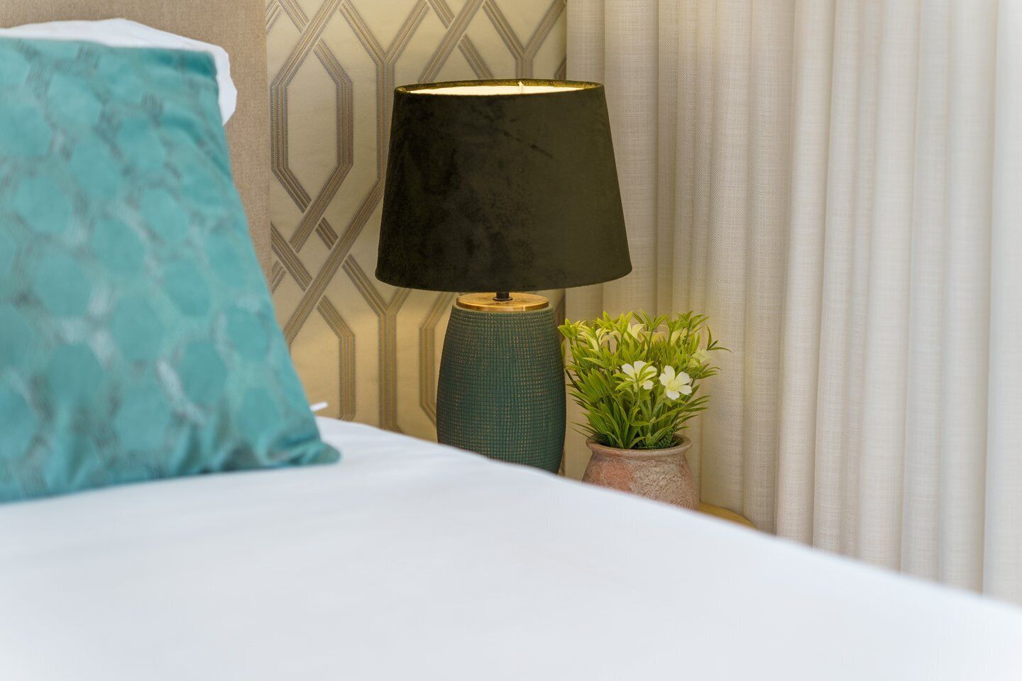A neatly made bed with a teal pillow, a dark lamp with a gold base, and a small potted plant with white flowers.