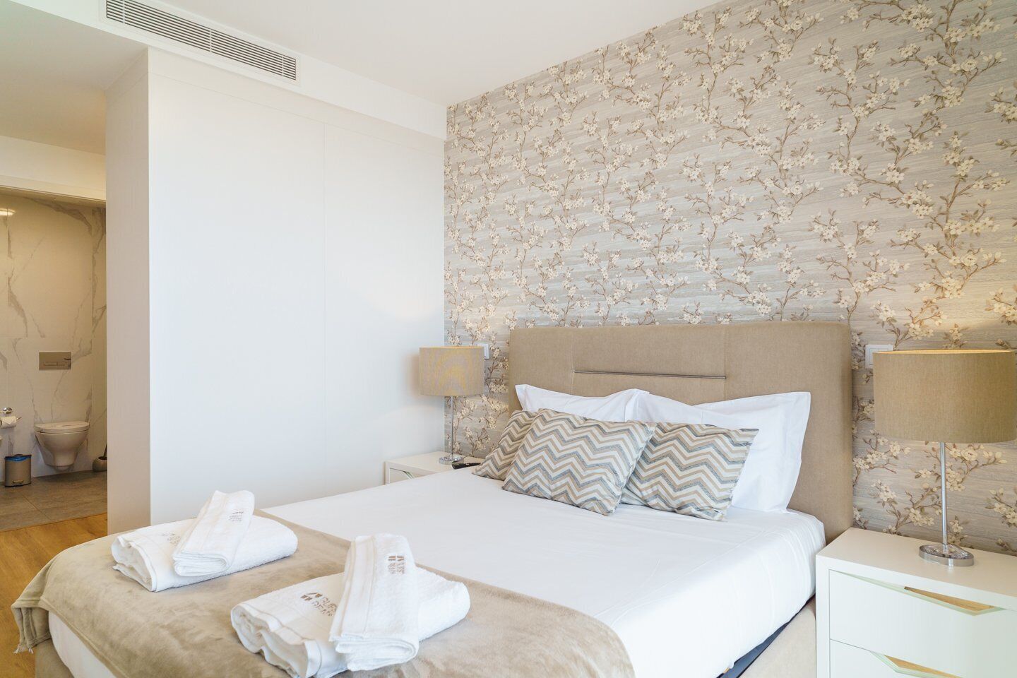 A hotel room with a bed, two pillows, a nightstand, a lamp, and a bathroom visible in the background.