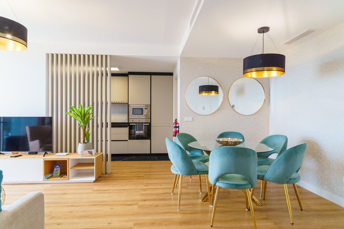 A modern living room with a TV, a dining area with teal chairs, and a kitchen in the background.